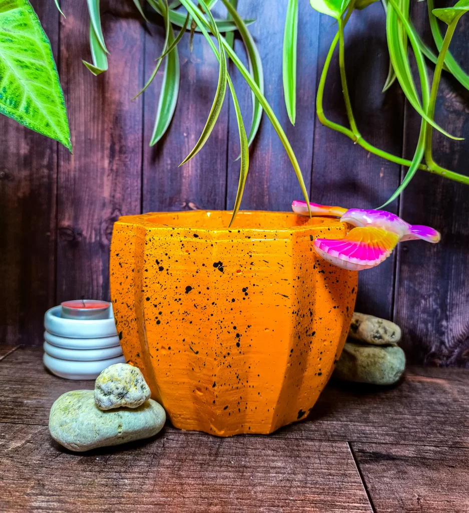 Maceta Zucca Naranja de concreto con forma de calabaza y 12 puntas, acabado jaspeado, en ambiente interior moderno con planta.