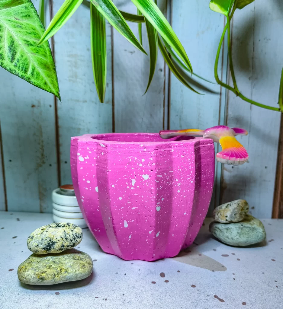 Maceta Zucca Fucsia de concreto con forma de calabaza y 12 puntas, acabado jaspeado, en ambiente interior moderno con planta.